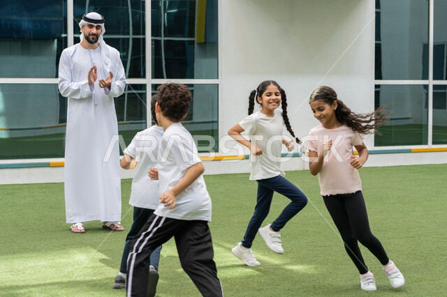 تطوير المهارة وتعزيز روح الفريق, ايماءات الفرح والسرور اثناء ممارسة التمارين الرياضية, تعليم الطلاب أساسيات الرياضة, مدرب عربي خليجي اماراتي يرتدي الغترة و الكندورة يشجع الأولاد بتعابير التصفيق, قضاء وقت ممتع في مدارس الامارات العربية المتحدة