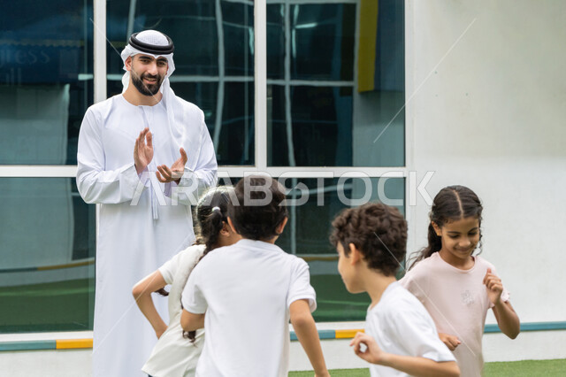 ايماءات الفرح والسرور اثناء ممارسة التمارين الرياضية, تعليم الطلاب أساسيات الرياضة, تطوير المهارة وتعزيز روح الفريق, مدرب عربي خليجي اماراتي يرتدي الغترة و الكندورة يشجع الأولاد بتعابير التصفيق, قضاء وقت ممتع في مدارس الامارات العربية المتحدة
