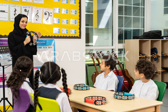 تنمية المهارة الموسيقية، قضاء وقت ممتع بالنشاط الترفيهي، تعلم استخدام الدف في المدرسة، معلمة عربية خليجية اماراتية محجبة ترتدي العباءة السوداء تقوم بالعزف على الآلات الإيقاعية، مجموعة من الطلاب يجلسون على المقاعد يحضرون درس الموسيقا بإيماءات التركيز
