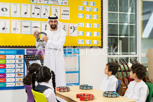 تعلم استخدام الدف في مدارس الامارات، معلم عربي خليجي اماراتي يرتدي الكندورة والغترة يقوم بالعزف على الآلات الإيقاعية، مجموعة من الطلاب يجلسون على المقاعد داخل الصف يحضرون درس الموسيقا بإيماءات التركيز، تنمية المهارة الموسيقية في المدرسة