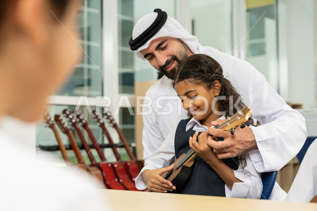 طالبة تجلس على المقعد داخل الصف تحاول استخدام الغيتار في حصة الموسيقى، مدرس عربي خليجي اماراتي يرتدي الكندورة والغترة يساعد تلميذته بالعزف على الآلات الوترية بتعابير السرور، تنمية الموهبة الموسيقية، قضاء وقت ممتع بالنشاط الترفيهي في المدرسة