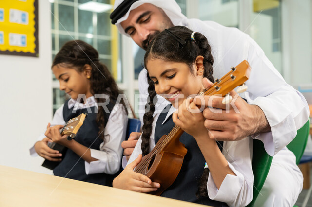 تنمية الموهبة الموسيقية، قضاء وقت ممتع بالنشاط الترفيهي في المدرسة، طالبتان تجلسان على المقعد داخل الصف تحاولان استخدام الغيتار في حصة الموسيقى، مدرس عربي خليجي اماراتي يرتدي الكندورة والغترة يساعد الاطفال بالعزف على الآلات الوترية بتعابير الفرح والسرور
