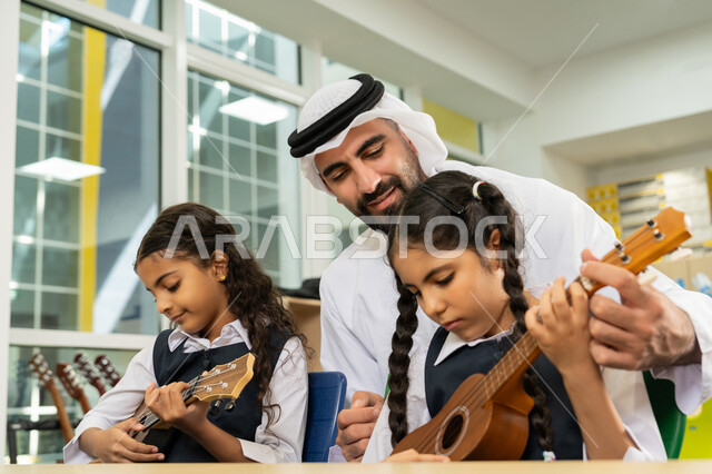 قضاء وقت ممتع بالنشاط الترفيهي في المدرسة، طالبتان تجلسان على المقعد داخل الصف تحاولان استخدام الغيتار في حصة الموسيقى، مدرس عربي خليجي اماراتي يرتدي الكندورة والغترة يساعد الاطفال بالعزف على الآلات الوترية بتعابير الفرح والسرور، تنمية الموهبة الموسيقية