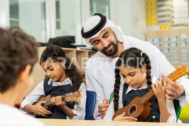 تعابير الفرح والسرور، طالبتان تجلسان على المقعد داخل الصف تحاولان استخدام الغيتار، مدرس عربي خليجي اماراتي يرتدي الكندورة والغترة يساعد الاطفال بالعزف على الآلات الوترية، تنمية الموهبة الموسيقية، قضاء وقت ممتع بالنشاط الترفيهي في المدرسة