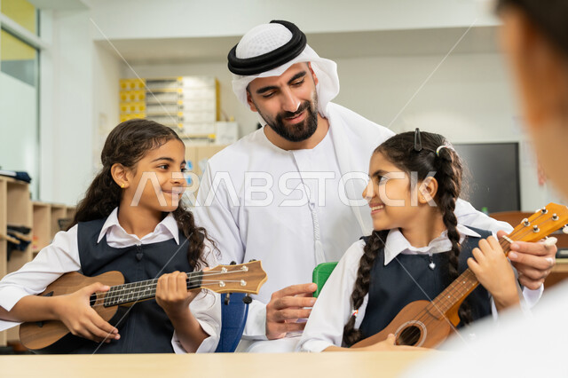 طالبتان تجلسان على المقعد داخل الصف تحاولان استخدام الغيتار، مدرس عربي خليجي اماراتي يرتدي الكندورة والغترة يساعد الاطفال بالعزف على الآلات الوترية في حصة الموسيقى، تنمية الموهبة الموسيقية، قضاء وقت ممتع بالنشاط الترفيهي في المدرسة