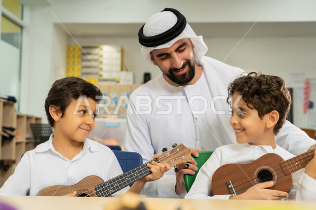 Developing musical talent, two students sitting on a bench inside the classroom trying to play the guitar with expressions of pleasure, having a good time in a recreational activity at school, an Arab Gulf Emirati teacher wearing a kandura and a ghutra helps children play stringed instruments in a music class