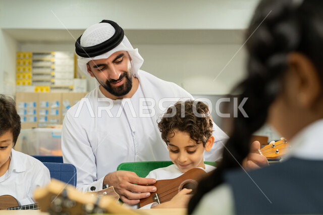 قضاء وقت ممتع بالنشاط الترفيهي في المدرسة، تنمية الموهبة الموسيقية، مدرس عربي خليجي اماراتي يرتدي الكندورة والغترة يساعد الاطفال بالعزف على الآلات الوترية بتعابير السرور، مجموعة من الطلاب يجلسون على المقاعد داخل الصف يحاولون استخدام الغيتار في حصة الموسيقى