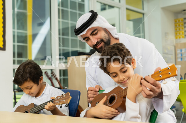 طالبان يجلسان على المقعد داخل الصف يحاولان استخدام الغيتار في المدرسة، قضاء وقت ممتع بالنشاط الترفيهي في مدارس الإمارات العربية المتحدة، مدرس عربي خليجي اماراتي يرتدي الكندورة والغترة يساعد الاطفال بالعزف على الآلات الوترية في حصة الموسيقى، تنمية الموهبة الموسيقية
