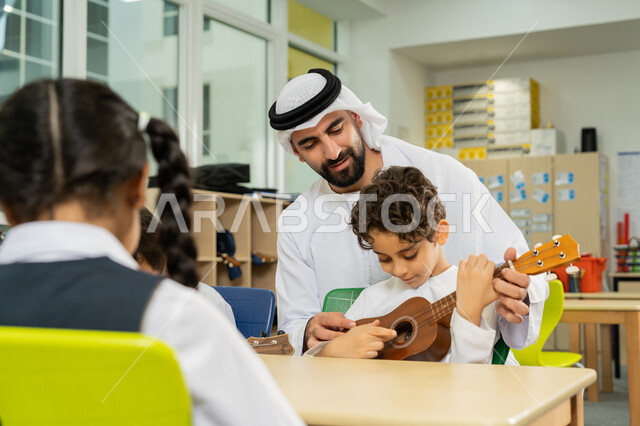 تنمية الموهبة الموسيقية في مدارس  الإمارات العربية المتحدة، مدرس عربي خليجي اماراتي يرتدي الكندورة والغترة يساعد الاطفال بالعزف على الآلات الوترية، مجموعة من الطلاب يجلسون على المقاعد داخل الصف يحاولون استخدام الغيتار، قضاء وقت ممتع بالنشاط الترفيهي في المدرسة