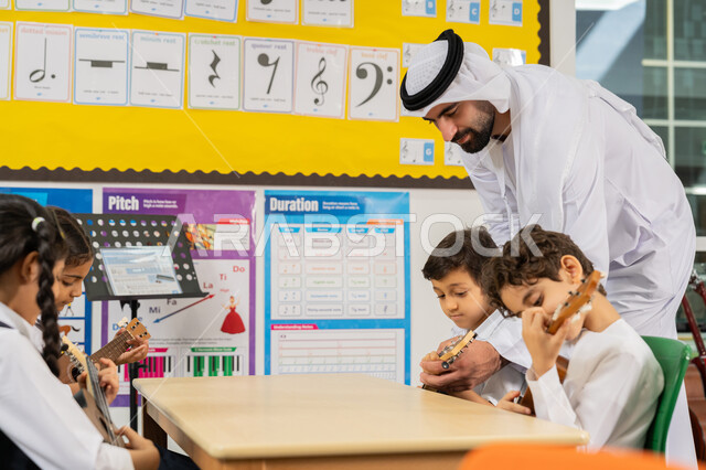 تنمية الموهبة الموسيقية، مدرس عربي خليجي اماراتي يرتدي الكندورة والغترة يساعد الاطفال بالعزف على الآلات الوترية، مجموعة من الطلاب يجلسون على المقاعد داخل الصف يحاولون استخدام الغيتار، قضاء وقت ممتع بالنشاط الترفيهي في المدرسة