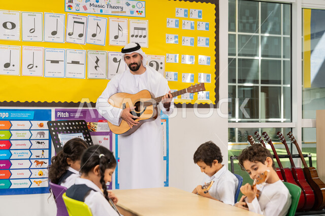 قضاء وقت ممتع في النشاط الترفيهي، معلم عربي خليجي اماراتي يرتدي الكندورة والغترة يقوم بالعزف على الآلات الوترية، تنمية الموهبة الموسيقية، مجموعة من الطلاب يجلسون على المقاعد داخل الصف يحضرون درس الموسيقا بإيماءات التركيز، تعلم استخدام الغيتار في المدرسة
