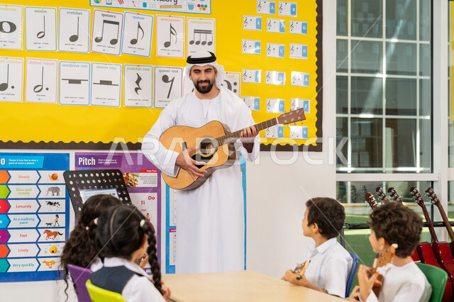 معلم عربي خليجي اماراتي يرتدي الكندورة والغترة يقوم بالعزف على الآلات الوترية، تنمية الموهبة الموسيقية، مجموعة من الطلاب يجلسون على المقاعد داخل الصف يحضرون درس الموسيقا بإيماءات التركيز، قضاء وقت ممتع في النشاط الترفيهي، تعلم استخدام الغيتار في المدرسة