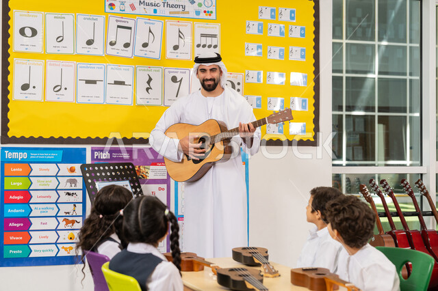 تنمية الموهبة الموسيقية، معلم عربي خليجي اماراتي يرتدي الكندورة والغترة يقوم بالعزف على الآلات الوترية، مجموعة من الطلاب يجلسون على المقاعد داخل الصف يحضرون درس الموسيقا بإيماءات التركيز، قضاء وقت ممتع في النشاط الترفيهي، تعلم استخدام الغيتار في المدرسة