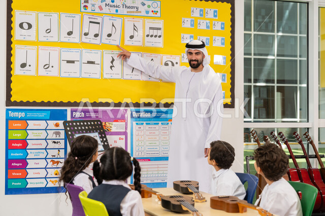Having fun in a recreational activity in the United Arab Emirates, learning to play guitar at school, an Emirati Gulf Arab teacher wearing a kandura and ghutra explaining musical notations, a group of students sitting on chairs inside the classroom attending a music lesson with gestures of concentration