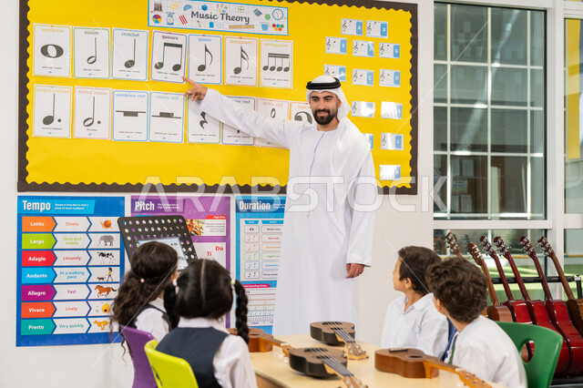 تعلم استخدام الغيتار في المدرسة، معلم عربي خليجي اماراتي يرتدي الكندورة والغترة يقوم بشرح العلامات الموسيقية، مجموعة من الطلاب يجلسون على المقاعد داخل الصف يحضرون درس الموسيقا بإيماءات التركيز، قضاء وقت ممتع في النشاط الترفيهي في الامارات العربية المتحدة