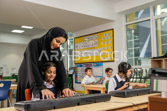 قضاء وقت ممتع في النشاط الترفيهي في مدارس الإمارات، معلمة عربية خليجية اماراتية محجبة ترتدي العباءة تقوم بمساعدة تلميذتها في استخدام الآلات الموسيقية، مجموعة من الطلاب يجلسون على المقاعد داخل الصف يضعون سماعات الرأس يقومون بالعزف على البيانو في حصة الموسيقى