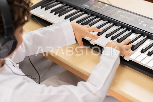 تنمية الموهبة والقدرة الموسيقية في الإمارات العربية المتحدة، صورة مقربة لولد عربي خليجي اماراتي يضع سماعات الرأس يقوم بتعلم العزف على آلة البيانو في حصة الموسيقى، قضاء وقت ممتع بالنشاط الترفيهي، ممارسة الهواية المفضلة