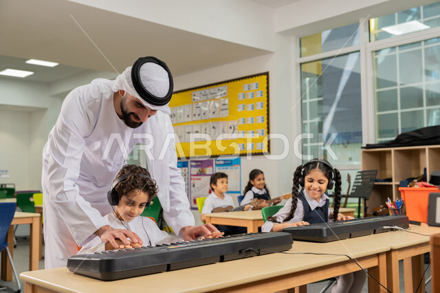 قضاء وقت ممتع بالنشاط الترفيهي في مدرسة في الإمارات، مدرس عربي خليجي اماراتي يرتدي الكندورة و الغترة يقوم بمساعدة التلميذ في استخدام الآلات الموسيقية، مجموعة من الطلاب يجلسون على المقاعد داخل الصف يضعون سماعات الرأس يقومون بالعزف على البيانو في حصة الموسيقى