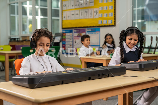 تنمية الموهبة والقدرة الموسيقية، مجموعة من الطلاب العرب الخليجيين الإماراتيين يجلسون على المقاعد داخل الصف يضعون سماعات الرأس يقومون بالعزف على البيانو في حصة الموسيقى، قضاء وقت ممتع في النشاط الترفيهي في مدارس الإمارات، تعلم استخدام الآلات الوترية