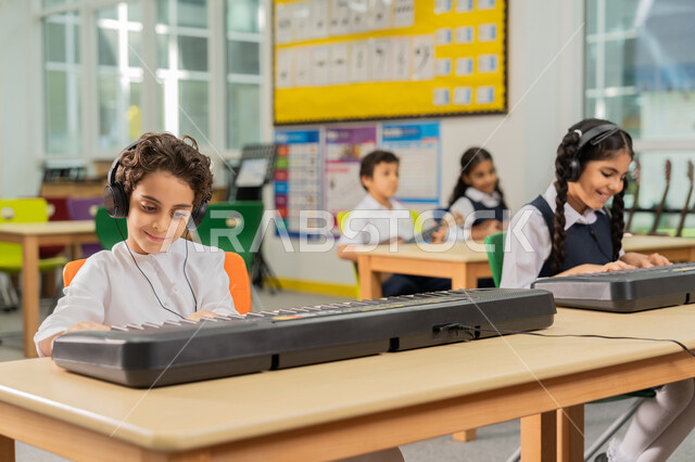 مجموعة من الطلاب العرب الخليجيين الإماراتيين يجلسون على المقاعد داخل الصف يضعون سماعات الرأس يقومون بالعزف على البيانو في حصة الموسيقى، قضاء وقت ممتع في النشاط الترفيهي في مدارس الإمارات، تعلم استخدام الآلات الوترية، تنمية الموهبة والقدرة الموسيقية