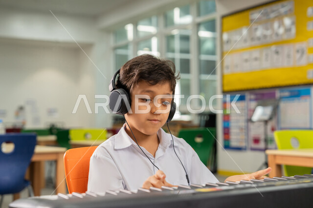 تنمية الموهبة والقدرة الموسيقية، صورة مقربة لولد عربي خليجي اماراتي يجلس على المقعد داخل الصف يضع سماعات الرأس يقوم بتعلم العزف على آلة البيانو في حصة الموسيقى، قضاء وقت ممتع في النشاط الترفيهي في مدارس الإمارات العربية المتحدة