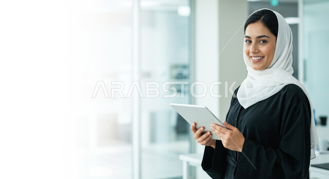 إدارة شؤون الأعمال وتنظيمها، استخدام الأجهزة التكنولوجية الحديثة والمتطورة، صورة مقربة لسيدة عربية خليجية سعودية محجبة مبتسمة تنظر الى الكاميرا وتحمل بيدها جهاز التابلت، العمل لتحقيق الاهداف واستراتيجيات الشركة، مهن ووظائف ادارية مكتبية