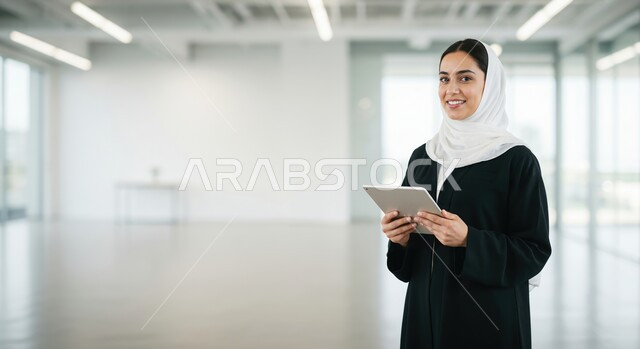Using modern and advanced technological devices, managing and organizing business affairs, a smiling veiled Saudi Arabian Gulf woman looking at the camera and holding a tablet in her hand, working to achieve the company's goals and strategies, administrative office jobs and professions