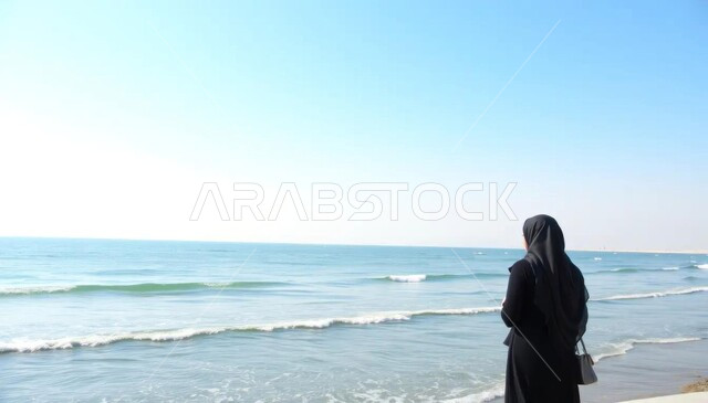 الحاجة إلى التأمل والاسترخاء أمام البحر وقت النهار، الشعور بالاكتئاب والوحدة، الاستمتاع بأوقات الفراغ في المناطق الساحليه السياحيه, صورة من الخلف لامراة عربية خليجية سعودية محجبة تقف امام البحر