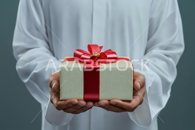 التعبير عن المحبة والتقدير, تقديم الهدايا في المناسبات السعيدة, الاحتفال بأعياد الميلاد, بورتريه مقرب لرجل عربي خليجي سعودي يرتدي الثوب التقليدي يمسك بيديه علبة هدية, خلفية رمادية