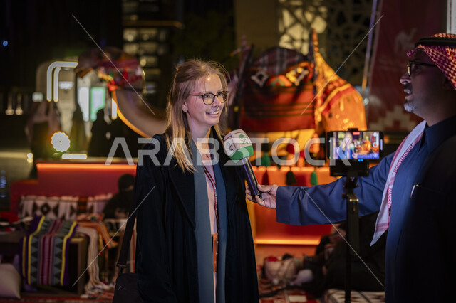 إجراء المقابلات مع السياح الأجانب، مذيع عربي خليجي سعودي يرتدي الشماغ والثوب التقليدي يمسك بيده مكبر الصوت يجري مقابلة مع سائحة اجنبية في مهرجان عام الابل في الرياض، تعزيز الهوية الثقافية وترسيخ التراث السعودي، أنشطة عربية شعبية تراثية