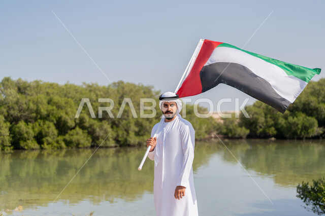 UAE National Day 1971 Remembrance, Union Founding Anniversary December 2, Emirati Gulf Arab Man Wearing Kandora and White Ghutra Holding UAE Flag Standing in Front of Pool of Water and Green Trees, Flag Day November 3, Love and Pride of the Nation, Nature Background