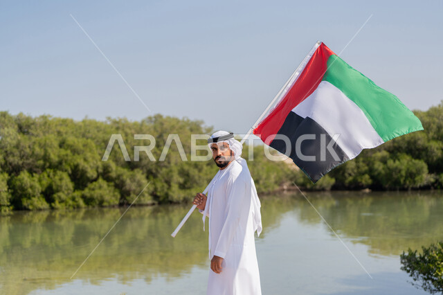 يوم العلم 3 نوفمبر، حب الوطن والاعتزاز به، استذكار اليوم الوطني الاماراتي 1971، ذكرى تأسيس الاتحاد  2 ديسمبر، رجل عربي خليجي إماراتي يرتدي الكندورة والغترة البيضاء يمسك بيده علم الإمارات العربية المتحدة يقف امام بركة المياه والاشجار الخضراء، خلفية الطبيعة
