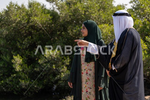 Pointing to something, hiking in natural areas, celebrating the anniversary of the founding of the union on December 2, love and pride in the homeland, UAE National Day 1971, an Arab Gulf Emirati man wearing a kandura and a bisht with a ghutra standing with a woman wearing an abaya and hijab in front of a large green tree during the day
