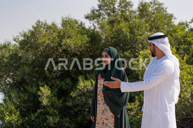 تبادل الاحاديث والتنزه في المناطق الطبيعية، الاحتفال بذكرى تأسيس الاتحاد 2 ديسمبر، حب الوطن والاعتزاز به، اليوم الوطني الاماراتي 1971، شاب عربي خليجي إماراتي يرتدي الكندورة والغترة يقف مع إمراة ترتدي العباءة والحجاب امام شجرة خضراء كبيرة في النهار