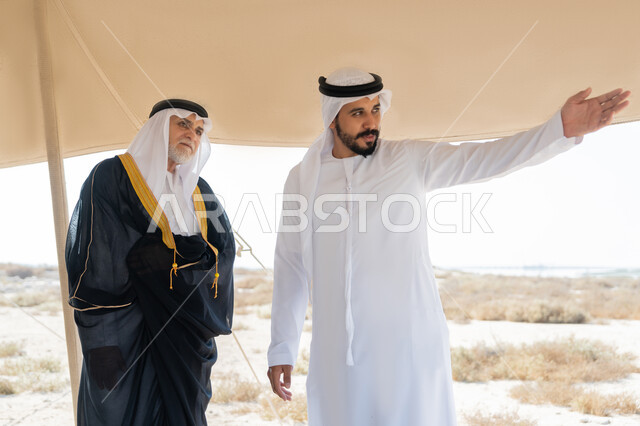 Pointing to something, Union anniversary December 2, UAE National Day celebration 1971, Close-up of a young Emirati Gulf Arab man wearing a white kandura and ghutra standing with a man wearing a bisht inside a tent in the desert, exchanging conversations with gestures of happiness, pleasure and pride