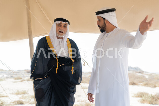 الاشارة الى شيء ما، ذكرى تأسيس الاتحاد 2 ديسمبر، الاحتفال باليوم الوطني الاماراتي 1971، صورة مقربة لشاب عربي خليجي إماراتي يرتدي الكندورة والغترة البيضاء يقف مع رجل يرتدي البشت داخل خيمة في الصحراء، تبادل الاحاديث بايماءات السعادة والسرور والاعتزاز