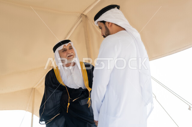 ذكرى تأسيس الاتحاد 2 ديسمبر، الاحتفال باليوم الوطني الاماراتي 1971، صورة مقربة لشاب عربي خليجي إماراتي يرتدي الكندورة والغترة البيضاء يقف مع رجل يرتدي البشت داخل خيمة في الصحراء، ايماءات السعادة والسرور والاعتزاز، تبادل الاحاديث وزيارة الاماكن التاريخية القديمة