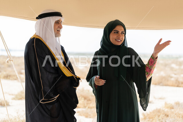 Exchanging conversations and visiting ancient historical places, the memory of the UAE National Day 1971, the founding of the union on December 2, a close-up photo of an Emirati Gulf Arab man wearing a kandura, ghutra and bisht standing with a woman wearing an abaya and hijab inside a tent in the desert, gestures of happiness, pleasure and pride