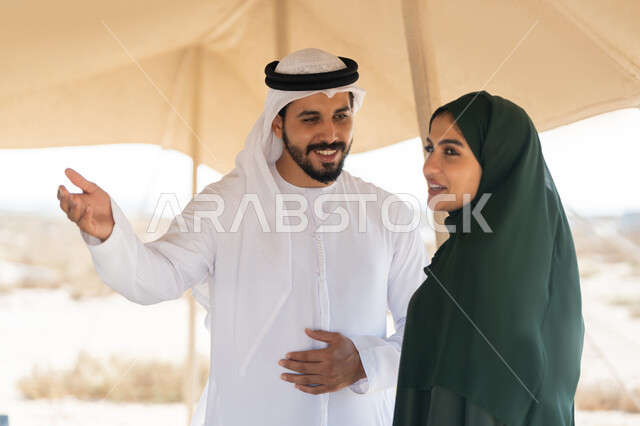 الاشارة الى شيء ما بتعابير السرور، تبادل الاحاديث وزيارة الاماكن التاريخية القديمة، ذكرى اليوم الوطني الاماراتي 1971، الاحتفال بتأسيس الاتحاد 2 ديسمبر، شاب عربي خليجي إماراتي يرتدي الكندورة والغترة البيضاء يقف مع إمراة ترتدي العباءة والحجاب داخل خيمة في الصحراء