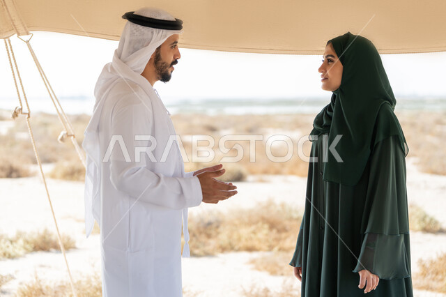 تعابير السعادة والسرور والاعتزاز، تبادل الاحاديث وزيارة الاماكن التاريخية القديمة، ذكرى اليوم الوطني الاماراتي 1971، الاحتفال بتأسيس الاتحاد 2 ديسمبر، شاب عربي خليجي إماراتي يرتدي الكندورة والغترة البيضاء يقف مع إمراة ترتدي العباءة والحجاب داخل خيمة في الصحراء