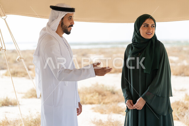 تبادل الاحاديث وزيارة الاماكن التاريخية القديمة، ذكرى اليوم الوطني الاماراتي 1971، الاحتفال بتأسيس الاتحاد 2 ديسمبر، شاب عربي خليجي إماراتي يرتدي الكندورة والغترة البيضاء يقف مع إمراة ترتدي العباءة والحجاب داخل خيمة في الصحراء، تعابير السعادة والسرور والاعتزاز