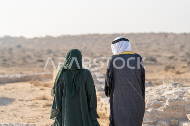 ايماءات الفخر والاعتزاز وحب الوطن، تبادل الاحاديث وزيارة الاماكن التاريخية القديمة، اليوم الوطني الاماراتي 1971، تأسيس الاتحاد 2 ديسمبر، صورة من الخلف لرجل عربي خليجي إماراتي يرتدي الكندورة والغترة والبشت يتجول مع إمراة ترتدي العباءة والحجاب في الصحراء الاماراتية