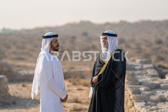 Wearing traditional costumes on national occasions, exchanging conversations and visiting historical places, UAE National Day 1971, the founding of the union on December 2, a young Emirati Gulf Arab man wearing a kandura and a ghutra walking with a man wearing a bisht in an archaeological area in the desert, pride, honor and love of the homeland