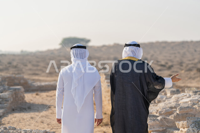 الاشارة الى مكان ما، زيارة الاماكن التاريخية القديمة، اليوم الوطني الاماراتي 1971، تأسيس الاتحاد 2 ديسمبر، صورة من الخلف لشاب عربي خليجي إماراتي يرتدي الكندورة والغترة يتجول مع رجل يرتدي البشت في منطقة أثرية في الصحراء، الفخر والاعتزاز وحب الوطن