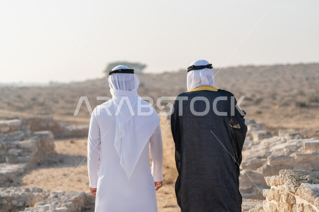 تبادل الاحاديث وزيارة الاماكن التاريخية القديمة، اليوم الوطني الاماراتي 1971، تأسيس الاتحاد 2 ديسمبر، صورة من الخلف لشاب عربي خليجي إماراتي يرتدي الكندورة والغترة يتجول مع رجل يرتدي البشت في منطقة أثرية في الصحراء، الفخر والاعتزاز وحب الوطن