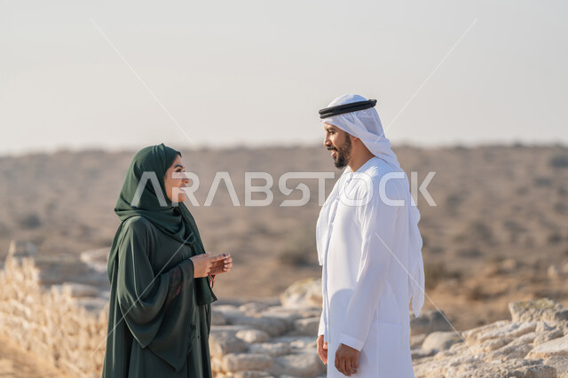 تبادل الاحاديث وزيارة الاماكن التاريخية القديمة، اليوم الوطني الاماراتي 1971، تأسيس الاتحاد 2 ديسمبر، شاب عربي خليجي إماراتي يرتدي الكندورة والغترة يتجول مع إمراة ترتدي العباءة والحجاب في منطقة أثرية في الصحراء، تعابير وايماءات الفخر والاعتزاز وحب الوطن