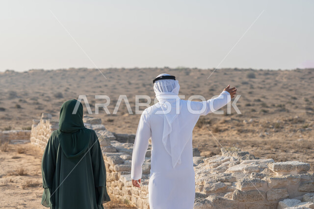 الفخر والاعتزاز وحب الوطن، الاشارة الى مكان ما، زيارة الاماكن التاريخية القديمة، اليوم الوطني الاماراتي 1971، تأسيس الاتحاد 2 ديسمبر، صورة من الخلف لشاب عربي خليجي إماراتي يرتدي الكندورة والغترة يتجول مع إمراة ترتدي العباءة والحجاب في منطقة أثرية في الصحراء