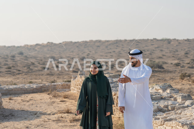 Exchanging conversations and visiting ancient historical places, UAE National Day 1971, the founding of the union on December 2, a young Emirati Gulf Arab man wearing a kandura and a ghutra walking with a woman wearing an abaya and hijab in an archaeological area in the desert, expressions and gestures of pride, honor and love of the homeland