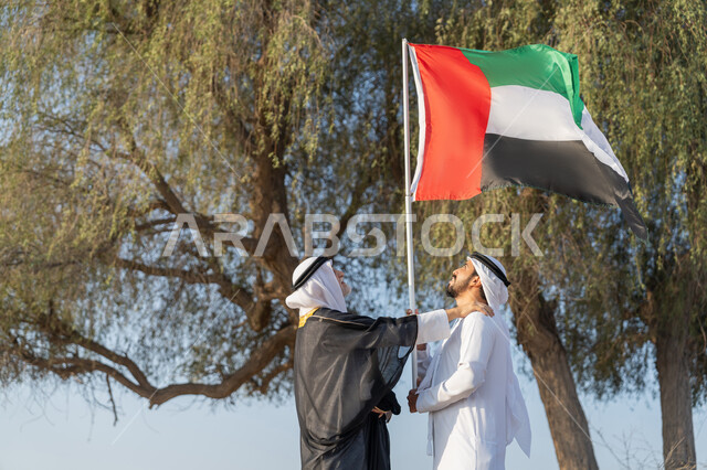 رجلان عربيان خليجيان اماراتيان يرتديان الزي التقليدي يرفعان علم الامارات العربية المتحدة في الصحراء، ارتداء الازياء الشعبية التقليدية للاحتفال بذكرى تأسيس الاتحاد 2 ديسمبر، يوم العلم 3 نوفمبر، حب الوطن والاعتزاز به، اليوم الوطني الاماراتي 1971، خلفية الطبيعة