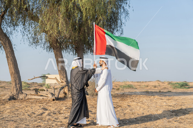 ارتداء الازياء الشعبية التقليدية للاحتفال بذكرى تأسيس الاتحاد 2 ديسمبر، يوم العلم 3 نوفمبر، حب الوطن والاعتزاز به، اليوم الوطني الاماراتي 1971، رجلان عربيان خليجيان اماراتيان يرتديان الزي التقليدي يرفعان علم الامارات العربية المتحدة في الصحراء، خلفية الطبيعة